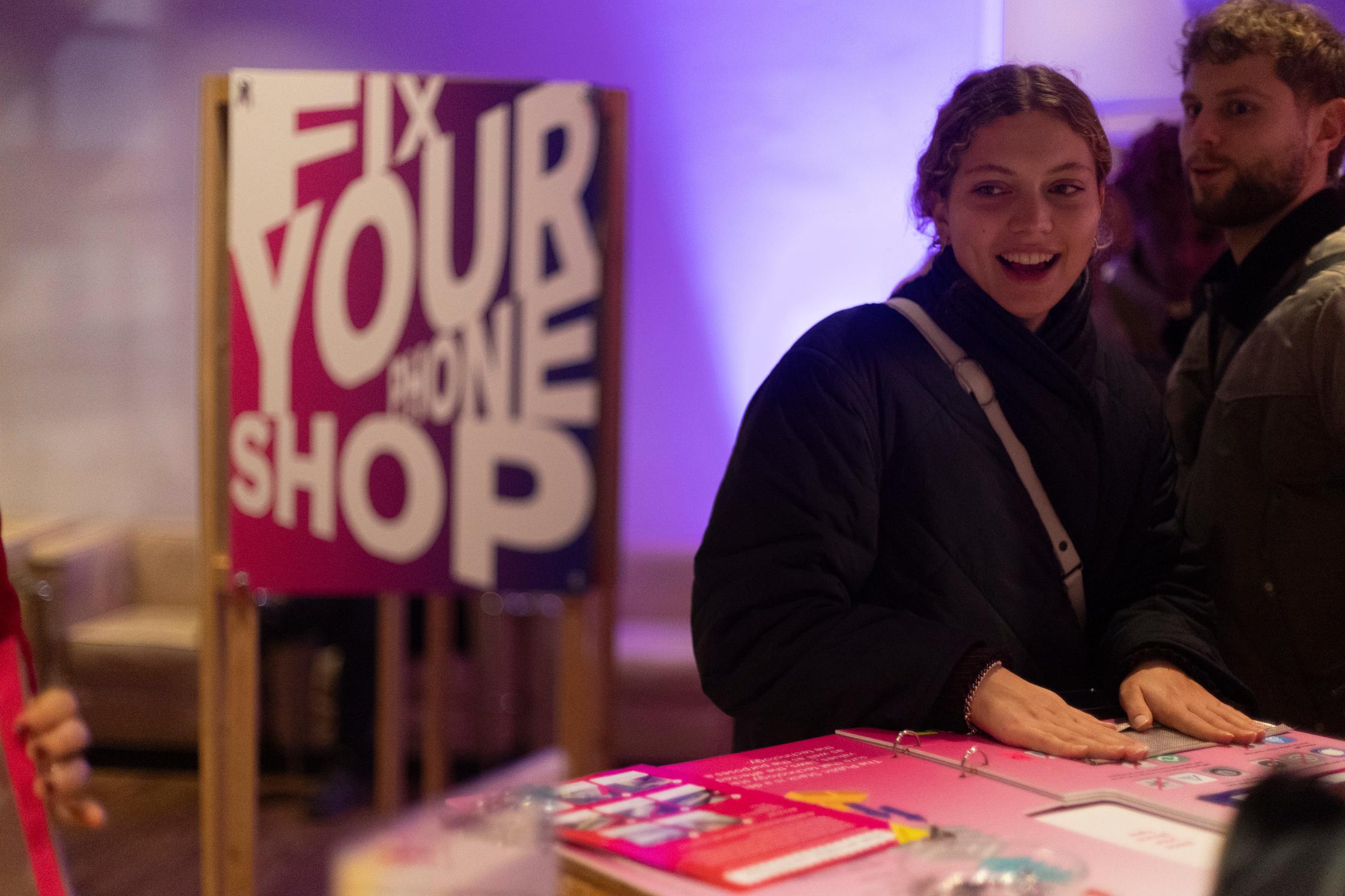 Een vrouw staat aan een tafel met daarop felgekleurde paarse flyers. Ze kijkt opzij. Achter haar staat een houten bord met 'Fix your phone shop' in grote witte letters tegen een rood-paarse achtergrond.