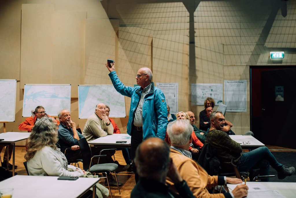 Een man staat in het midden van een zaal en houdt een sensor in de lucht. Om hem heen zitten mensen die naar hem kijken.