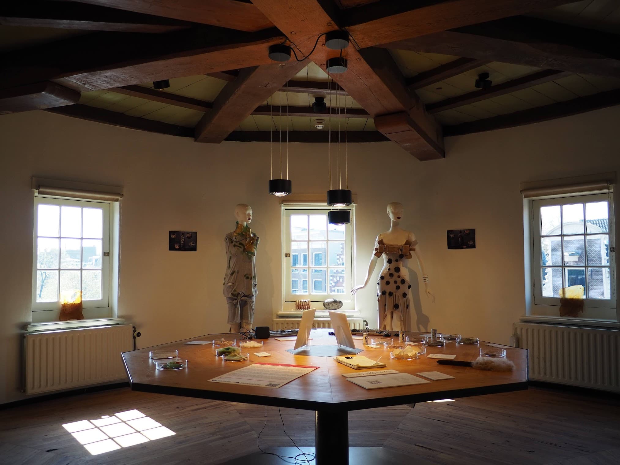 Photograph of a hexagonical shaped room in de Waag, with an installation on a hexagonical wooden table featuring two mannequins facing the viewer, and different textile materials stalled on the table