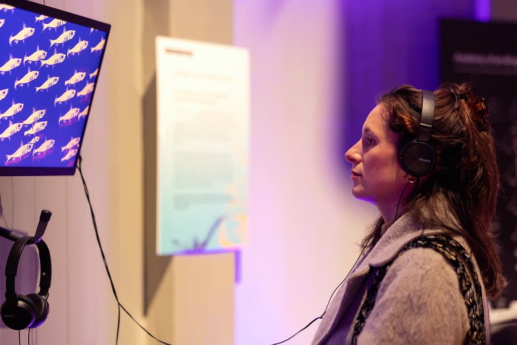 Foto van een bezoeker tijdens de museumnacht 2023 in Waag Futurelab. Een vrouw kijkt naar een beeldscherm, waarop lichtblauwe vissen te zien zijn tegen een donkerblauwe achtergrond. Ze heeft een hoofdtelefoon op.
