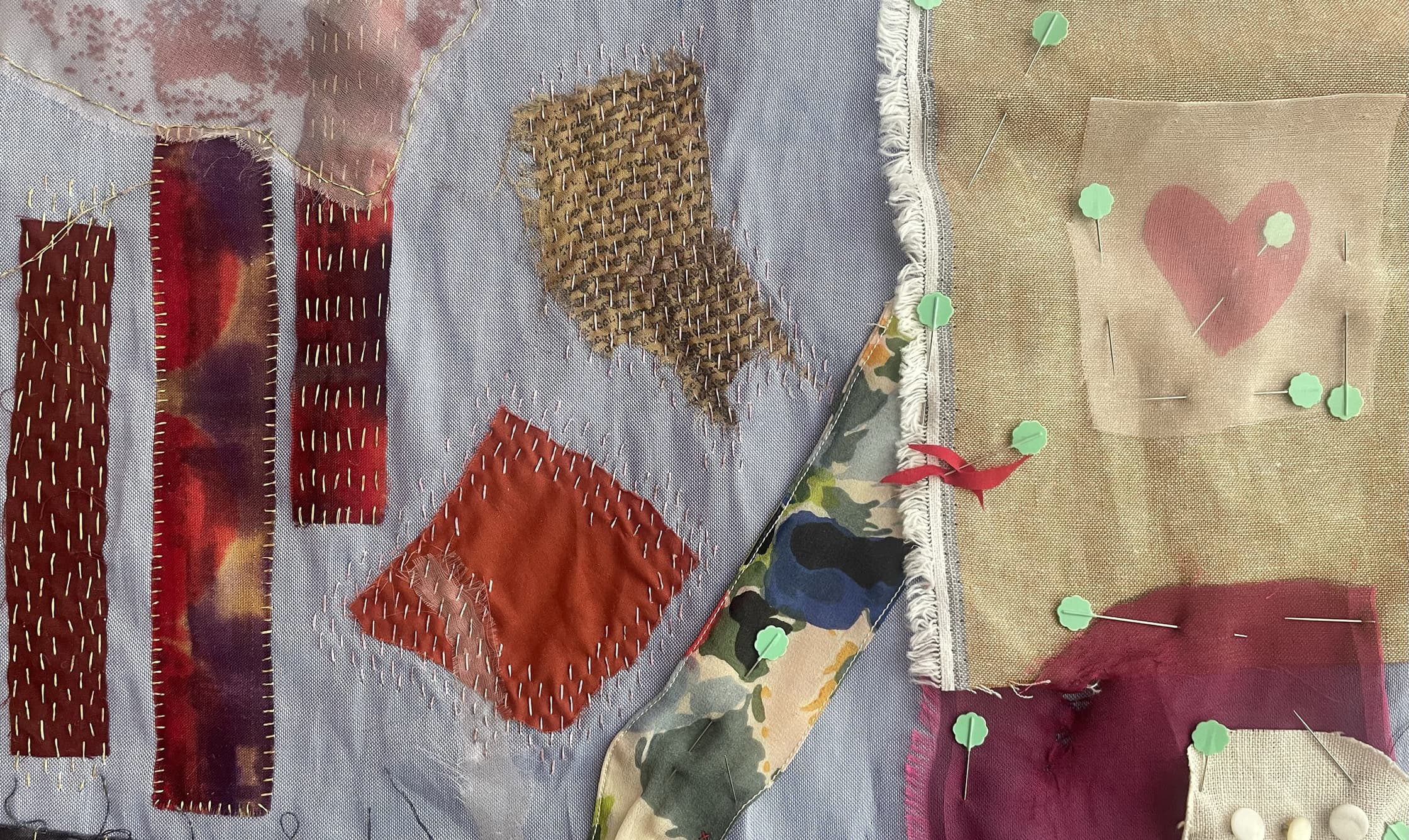 Een foto van geborduurde stiksels op een stuk wit linnen. Verschillende stukken stof zijn erop gestikt met kleurige stiksels in de kleuren bordeau rood, oranje, groen en lichtgeel. 