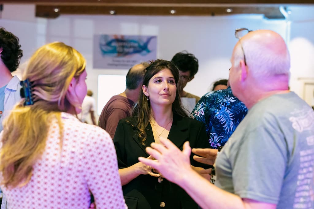 Drie mensen geëngageerd in gesprek tijdens een avond georganiseerd door de Groene Amsterdammer en Waag Futurelab. Een Vrouw met donker haar staat in het midden van de foto te luisteren naar een man die met zijn rug naar de camera toestaat. Links staat ook een vrouw met haar rug naar de camera.
