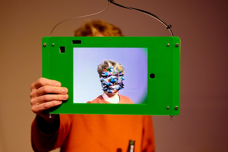 Man houdt een scherm voor zijn hoofd, waarop zijn hoofd wordt getoond maar dan met kleurige digitale vlakken op de plek van zijn gezicht.