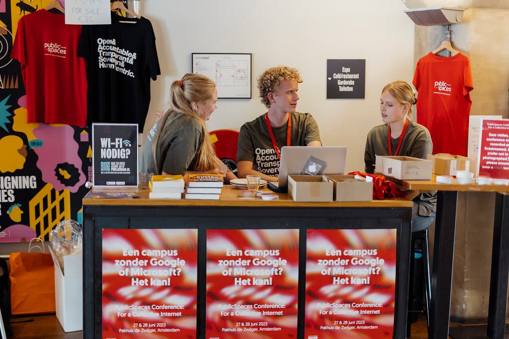 Drie mensen zitten achter een registratietafel bij de PublicSpaces conferentie. Aan de voorkant van de tafel hangen drie posters met de PublicSpaces vormgeving: rood met witte strepen. Een persoon in het middel zit achter een laptop en praat met de persoon aan diens linkerzijde.