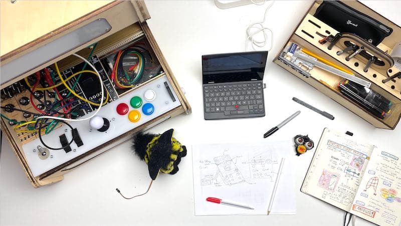 Picture of the work in progress of the Furby synthesiser module by Edwin Dertien. On the left there is a large wooden box opened with various electronic elements, such as big colorfol buttons, wires, switches and lights. Next to it there is a small laptop and a deconstructed Furby toy. There is also a technical drawing of the device and on the right a wooden toolbox like box with pens and other tools.