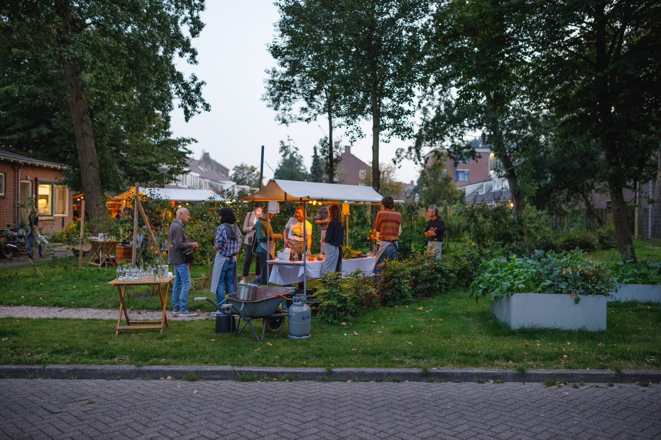 Een kraampje met sfeerverlichting. Rond het kraamje staan buurtbewoners met elkaar te praten. Het kraampje staat in het gras en is omringd door een paar bomen. Op de achtergrond zijn een aantal huizen te zien.